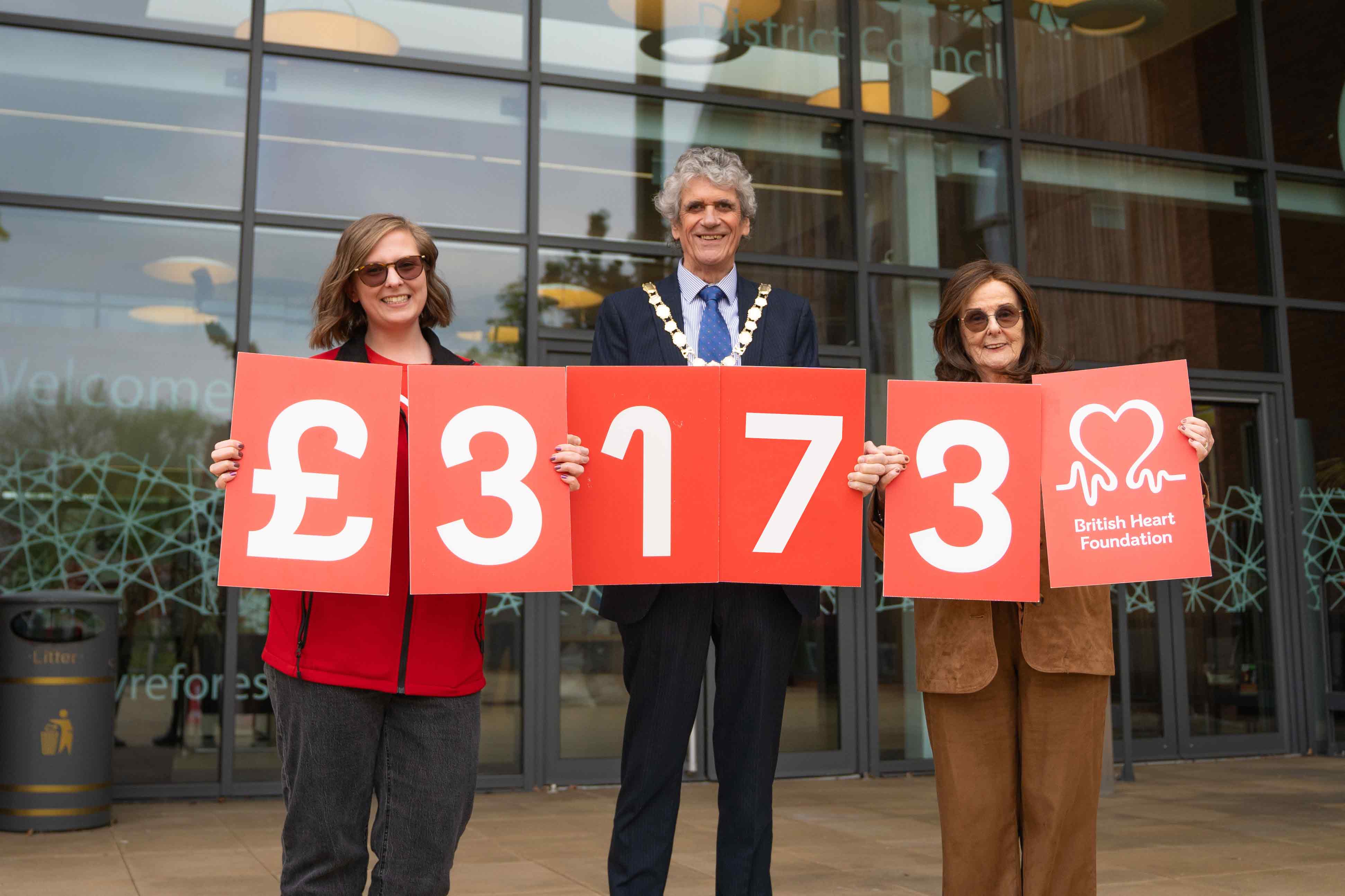 two females a male stood holding red signs with numbers on to represent how much money had been raised for the charity