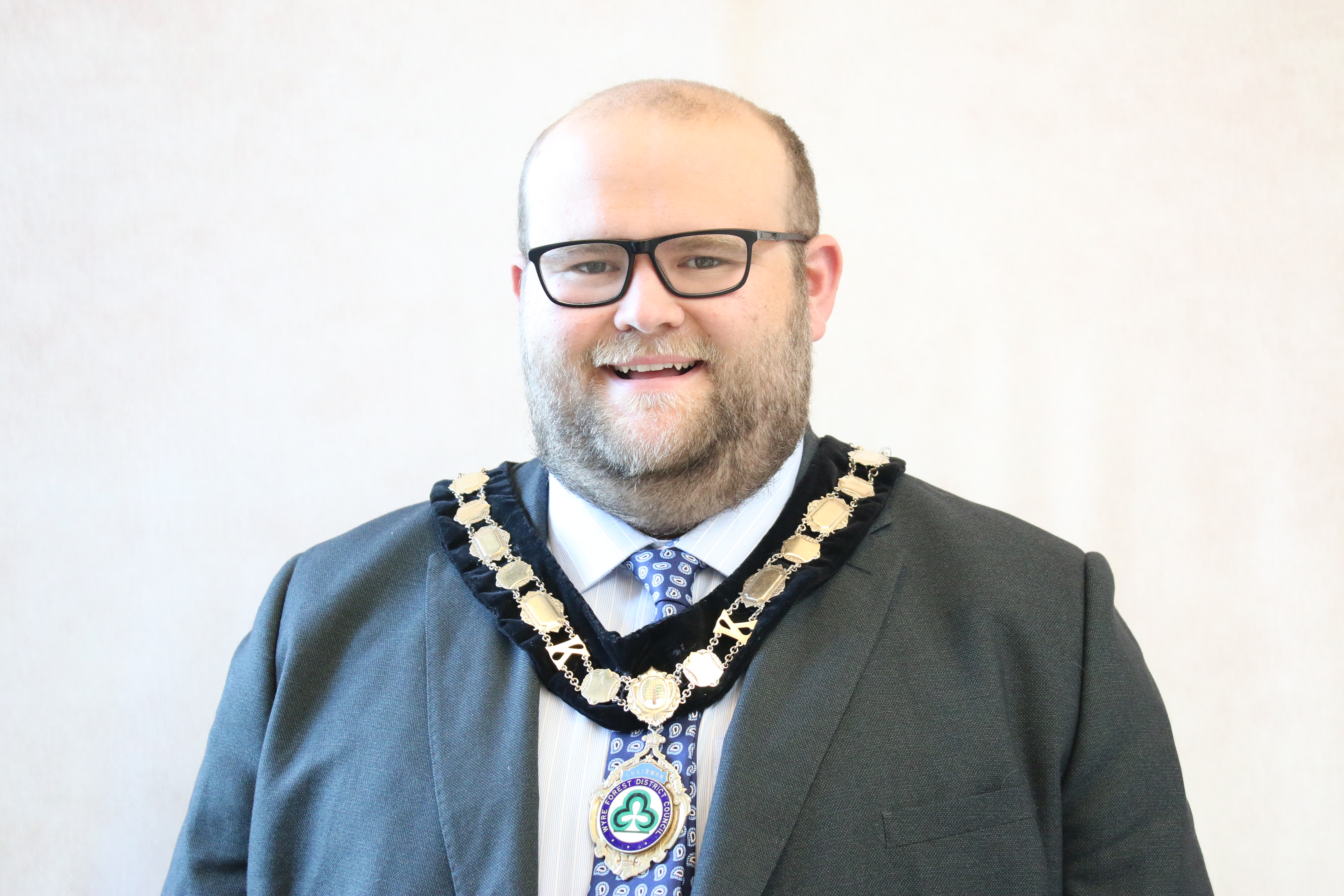 Male wearing a blue suit, white shirt and blue patterned  tie with Council chains on