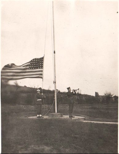 a soldier lowers US flag while another plays bugle at flag pole in field. Old sepia photo